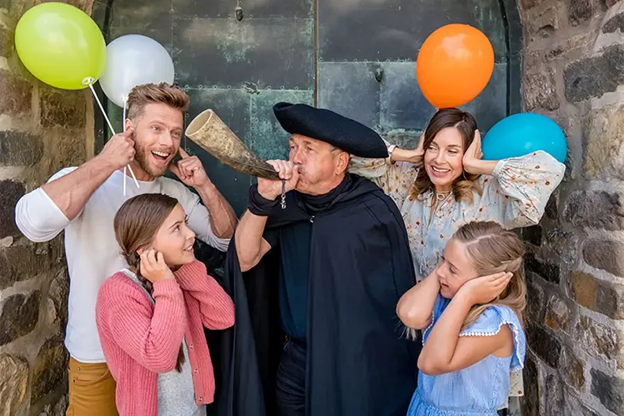 Der Nachtwaechter blaest vor einem alten Tor ins Signalhorn, eine Familie mit zwei Kindern haelt sich lachend die Ohren zu, bunte Luftballons ringsum