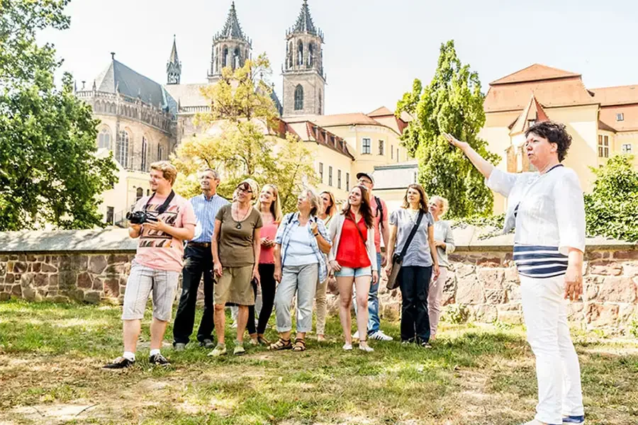 Stadtfuehrerin zeigt einer Besuchergruppe die Stadt: die Gruppe steht auf einer Wiese an der alten Festungsmauer und blickt auf, dahinter die Tuerme des Doms