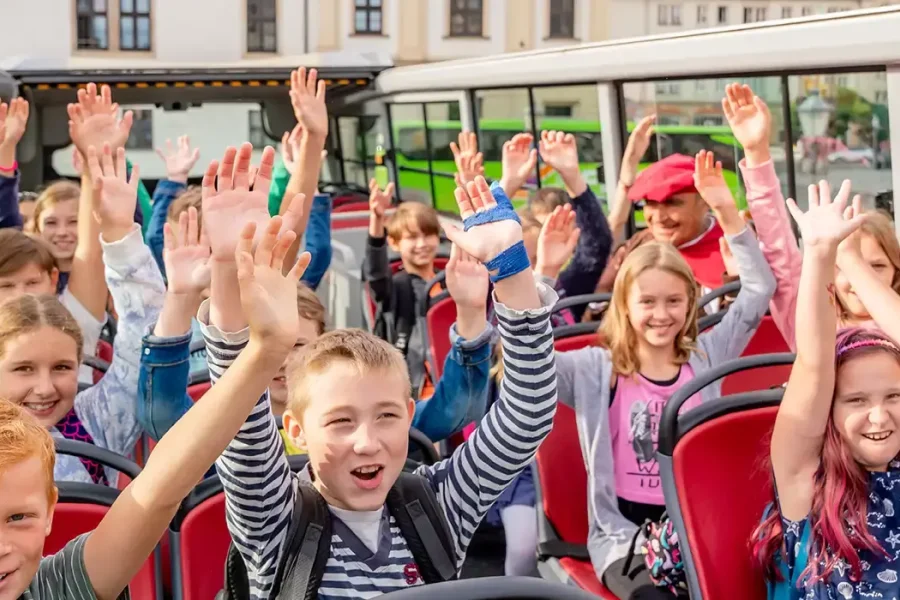 Kinder auf dem offenen Oberdeck des Doppeldeckerbusses strecken jubelnd die Arme hoch, im Hintergrund ein Denkmal und historische Fassaden