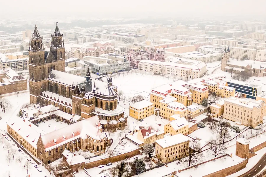Luftaufnahme des tief verschneiten Magdeburg: Dom und Klosterhoefe in Weiss, dahinter die Gruene Zitadelle und die Innenstadt im Winterdunst