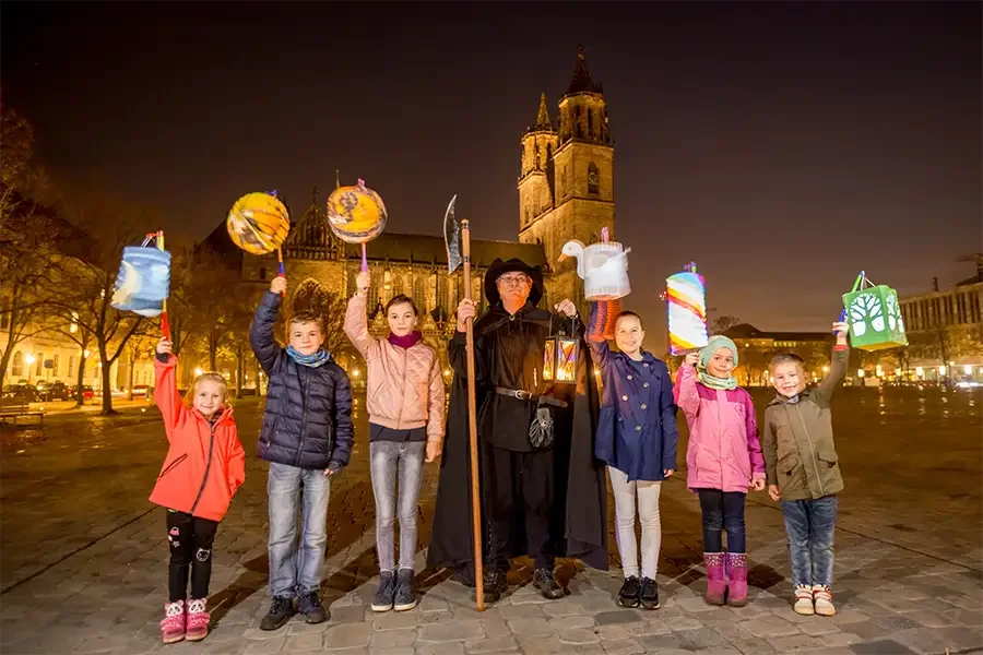 Nachtwaechter mit Hellebarde und Laterne umringt von Kindern mit bunten Lampions auf dem abendlichen Domplatz