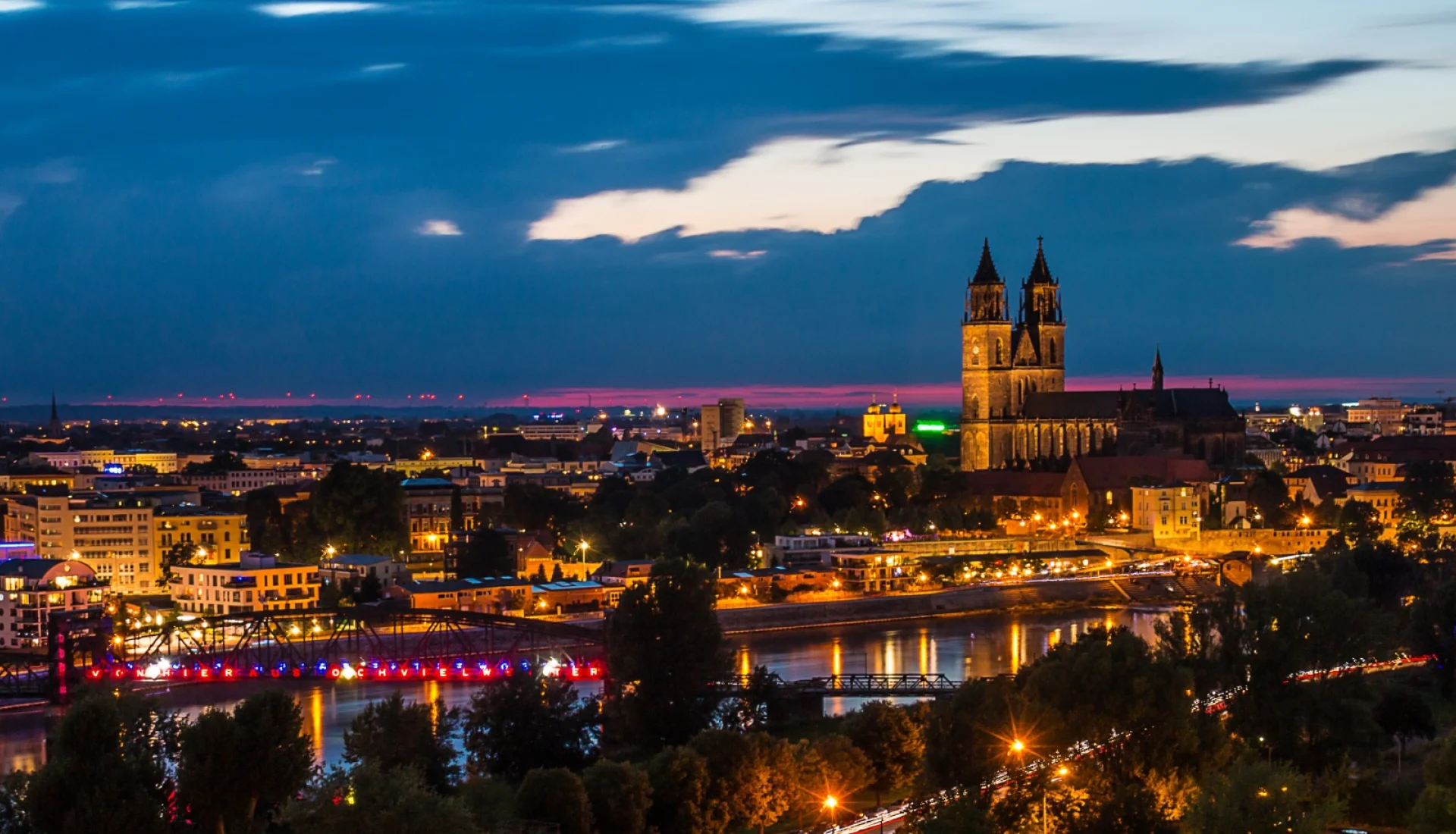 Naechtliches Panorama von Magdeburg: beleuchteter Dom ueber der Stadt, vorn die rote Leuchtschrift der alten Hubbruecke an der Elbe