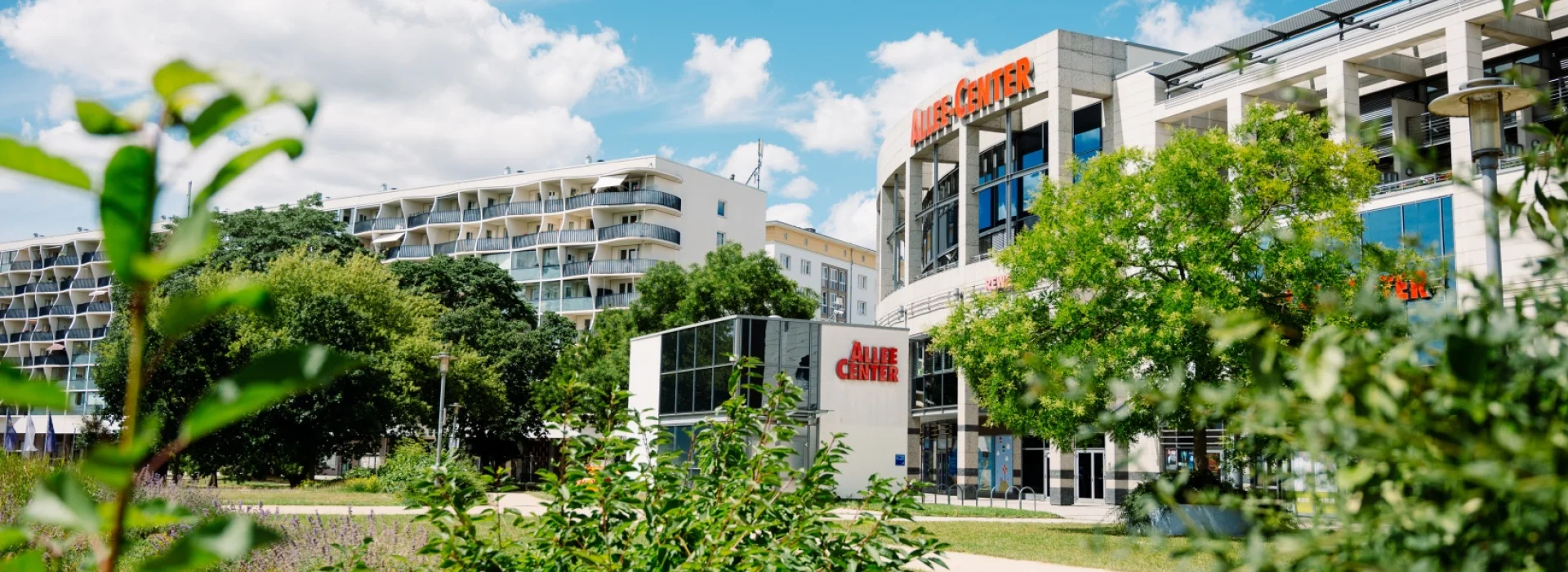 Allee-Center Magdeburg mit rotem Schriftzug &uuml;ber dem runden Eingangsbau, davor eine Wiese mit jungen Str&auml;uchern