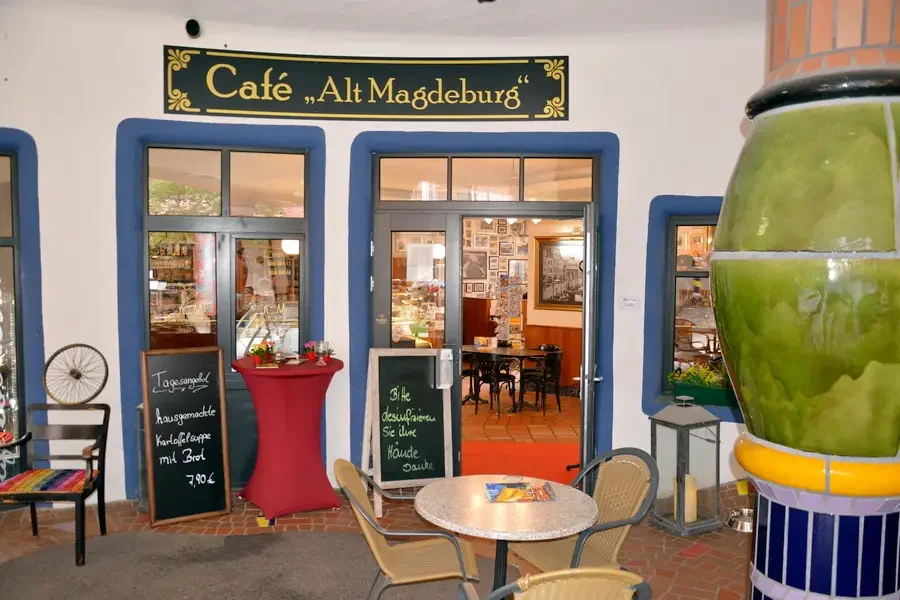 Eingang des Cafe Alt Magdeburg in der Gruenen Zitadelle: blau gerahmte Fenster, Angebotstafeln und Bistrotische, rechts eine gruene Keramiksaeule