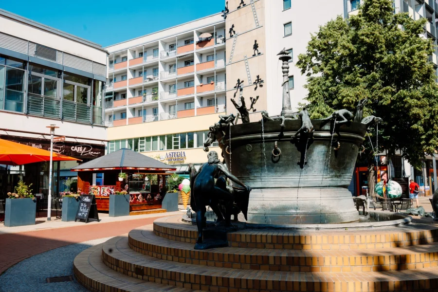 Faunenbrunnen mit seinen Bronzefiguren auf gestuftem Sockel, dahinter das Caf&eacute; Piano mit Terrasse und Sonnenschirmen