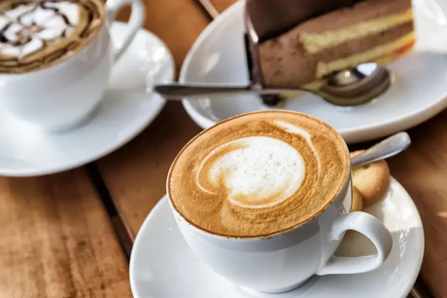 Cappuccino mit Milchschaum-Muster in weisser Tasse auf Holztisch, dahinter ein Stueck Schokoladentorte und eine zweite Kaffeetasse