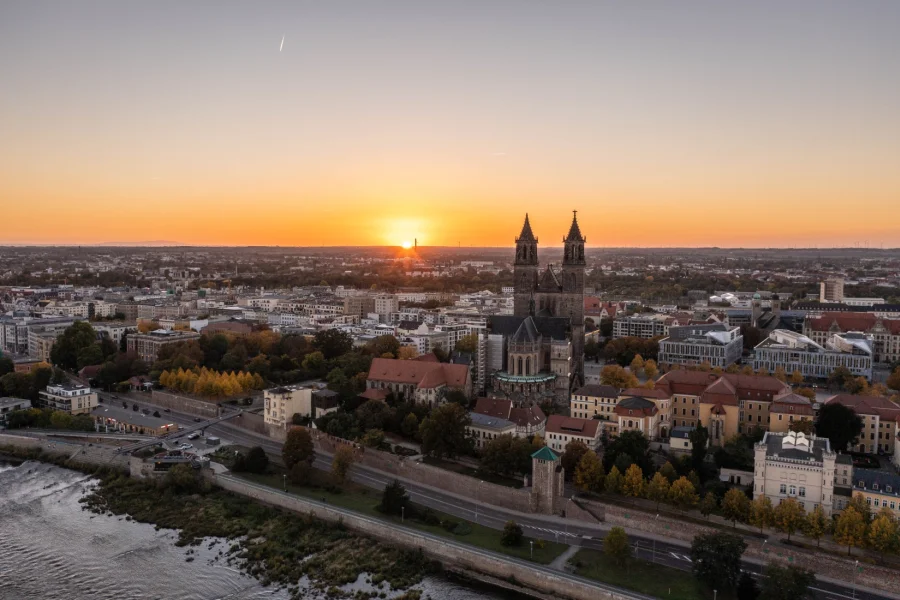Panorama von Magdeburg bei Sonnenuntergang: der Dom ueberragt die Stadt, vorn die Elbe mit Uferstrasse und Festungsmauer im Herbstlaub
