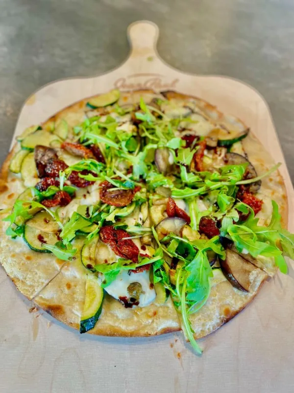 D&uuml;nner Flammkuchen mit Zucchini, Aubergine, getrockneten Tomaten, Mozzarella und Rucola auf einem hellen Holzschieber