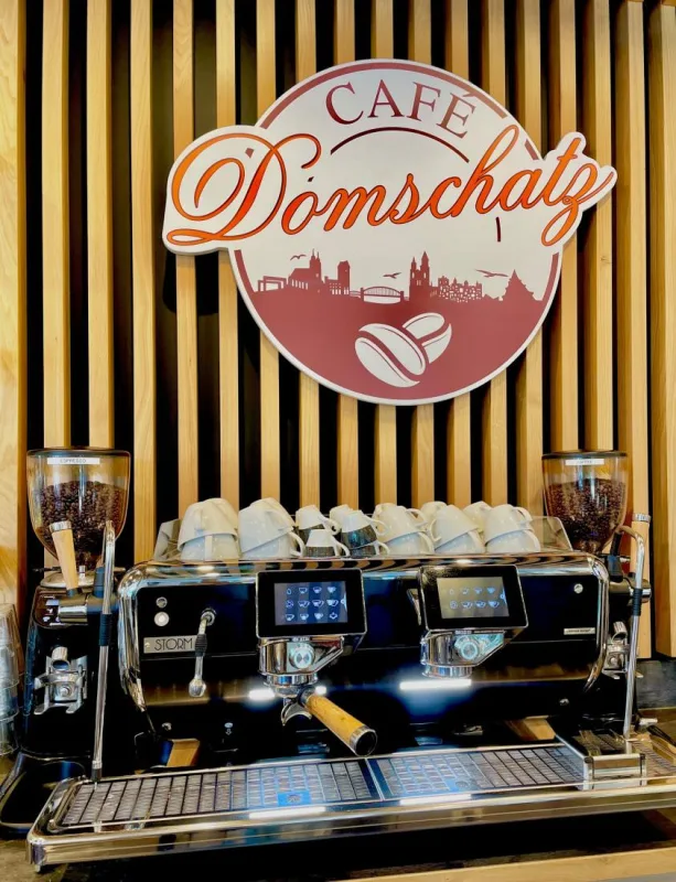 Siebtr&auml;germaschine mit vorgew&auml;rmten Tassen vor einer Holzlattenwand, dar&uuml;ber das Logo des Caf&eacute; Domschatz