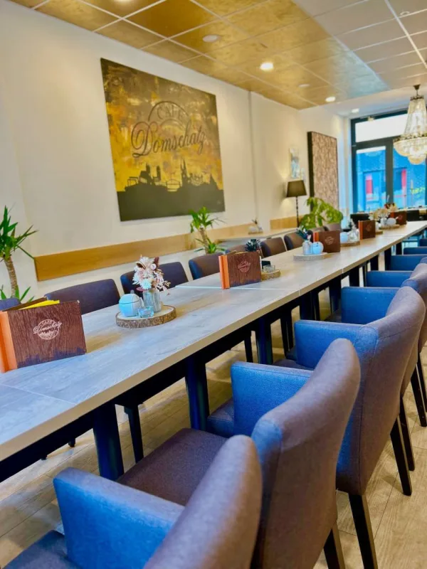 Lange Tafel mit blauen Polsterst&uuml;hlen im Gastraum des Caf&eacute; Domschatz, an der Wand ein goldfarbenes Bild mit dem Caf&eacute;-Schriftzug
