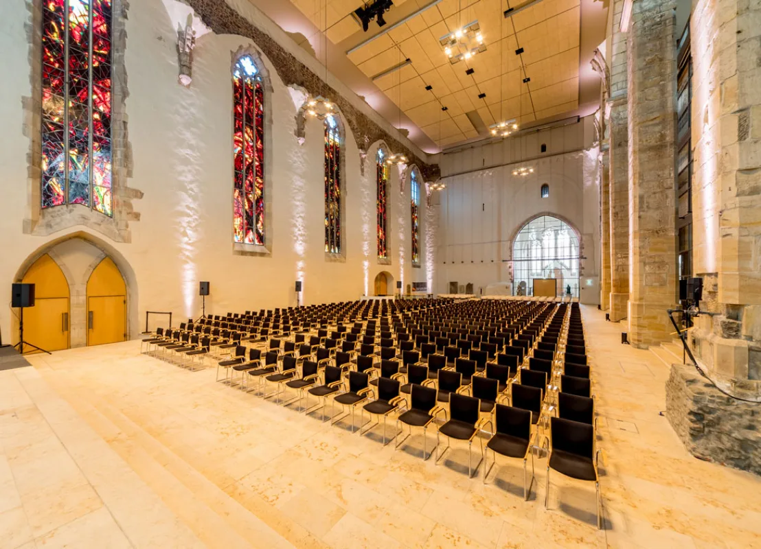 Kirchenschiff der Johanniskirche als Veranstaltungssaal: Reihenbestuhlung auf hellem Steinboden, bunte Kirchenfenster und Blick zur B&uuml;hne