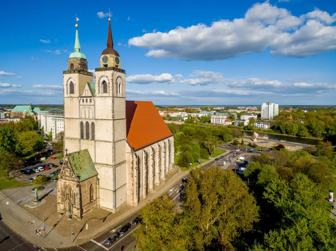 Luftbild der Johanniskirche mit rotem Dach und beiden T&uuml;rmen, dahinter die Elbe und die Magdeburger Innenstadt