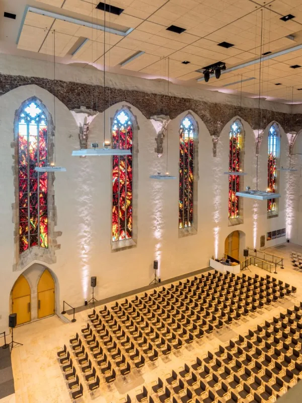Saal der Johanniskirche von der Empore aus: hohe gotische Fenster mit rot leuchtendem Glas, wei&szlig; verputzte W&auml;nde und dicht gestellte Stuhlreihen