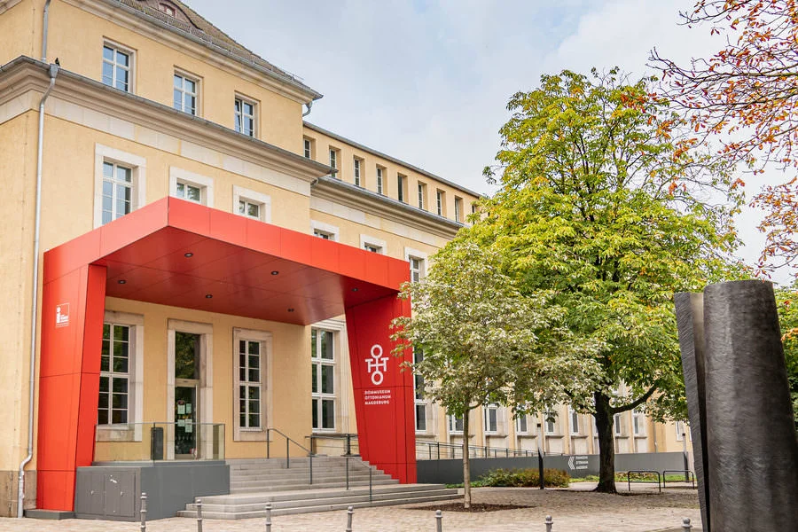 Roter Eingangsrahmen mit dem Signet des Dommuseums Ottonianum vor dem gelben Altbau, daneben eine junge Kastanie