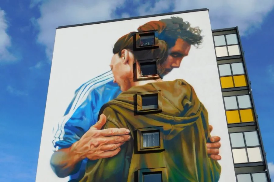 Gro&szlig;fl&auml;chiges Wandbild an einem Plattenbau: zwei Personen umarmen sich, das Motiv reicht &uuml;ber f&uuml;nf Fensterachsen, rechts eine Fassade mit gelben und wei&szlig;en Farbfeldern