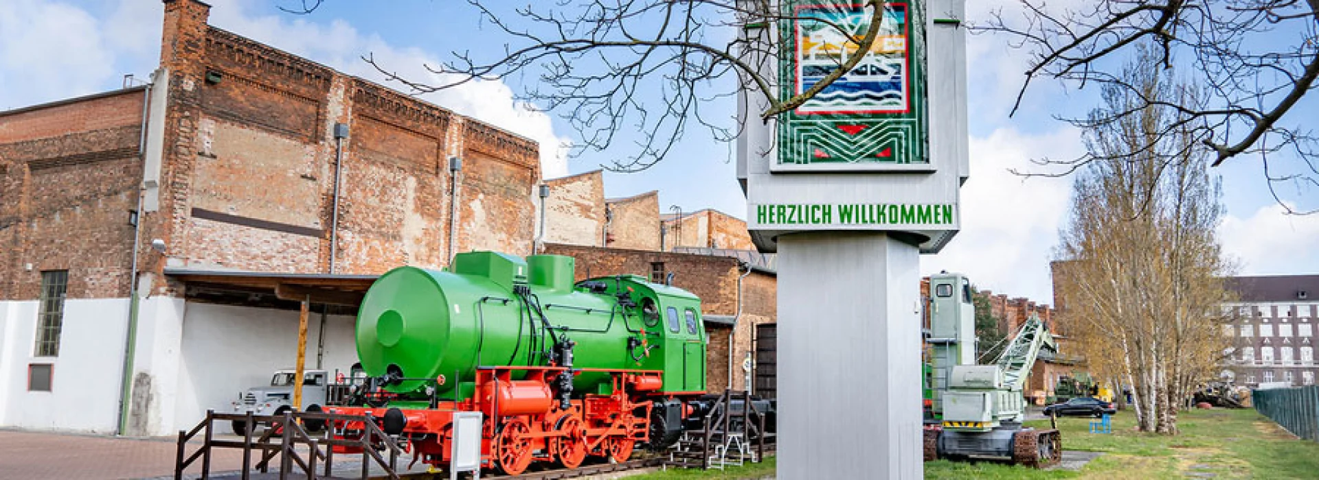 Gr&uuml;ne Dampflokomotive mit rotem Fahrwerk vor der Backsteinhalle des Technikmuseums, daneben eine Stele mit Buntglasfenster und der Aufschrift &bdquo;Herzlich willkommen"