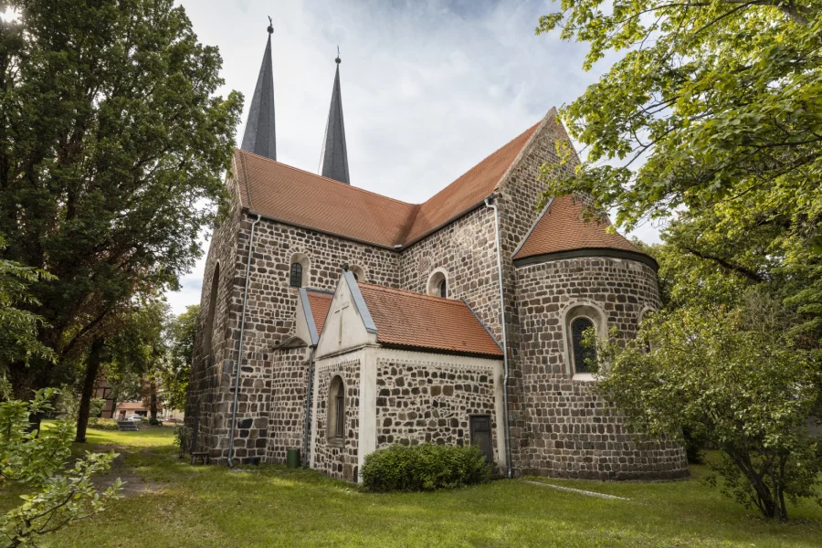 Die romanische Feldsteinkirche St. Nicolai in Burg: Bruchsteinmauerwerk mit roten Ziegeldaechern, halbrunder Apsis und zwei spitzen Turmhelmen zwischen alten Baeumen