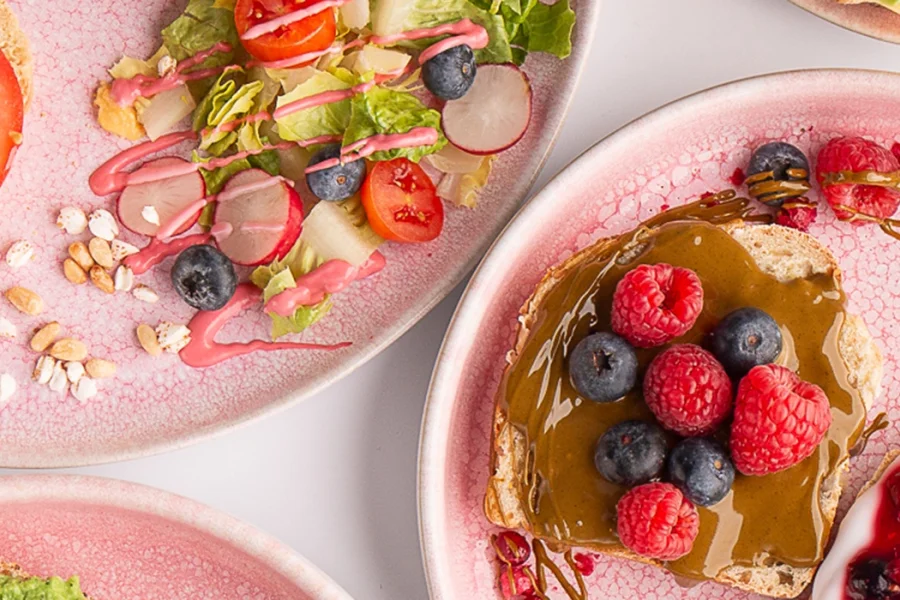 Fruehstuecksteller auf rosa Keramik im Vanillekind: Brote mit Tomate-Mozzarella-Basilikum, mit Nussmus und frischen Himbeeren und Heidelbeeren, dazu Salat mit Radieschen und Kirschtomaten