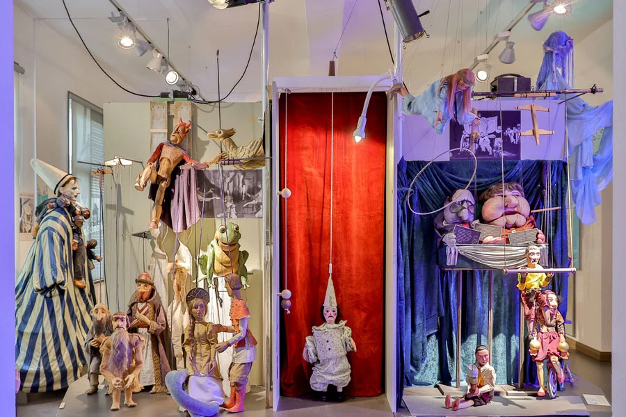 Ausstellungsraum der Figurenspielsammlung mit Marionetten und Stabfiguren vor einem roten B&uuml;hnenvorhang, rechts eine blau ausgeschlagene Puppenb&uuml;hne