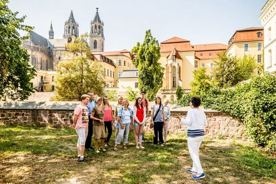 Stadtfuehrerin erklaert einer Gruppe von zehn Gaesten die Reste der Stadtmauer, im Hintergrund die beiden Tuerme des Magdeburger Doms